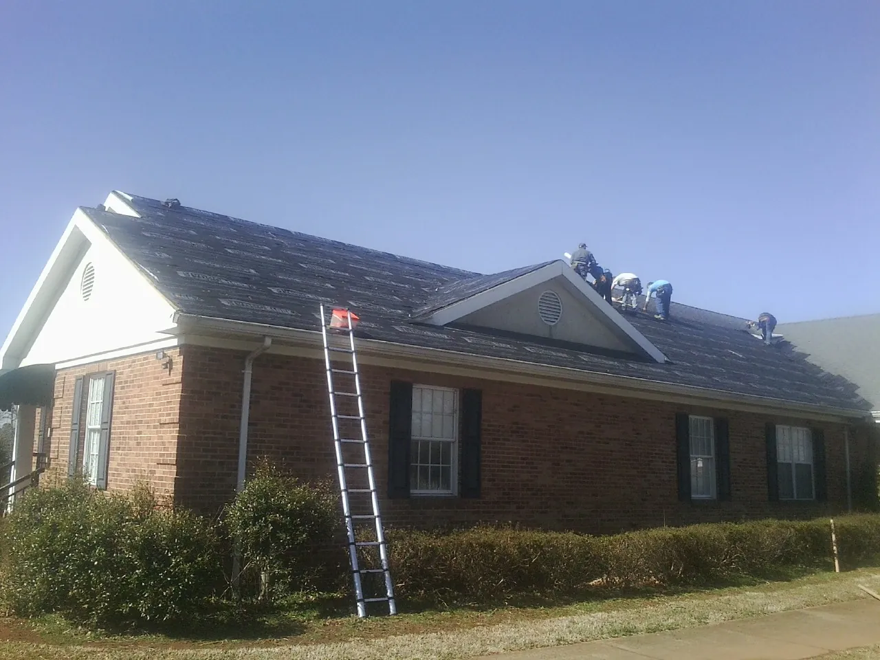Crew tearing shingles off a brick ranch roof with a ladder set — Metal Roof Repair work by our crew in New Boston